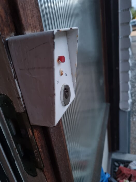 Vit, rektangulär låda monterad på dörrkarm med rött och grönt ljus, och en nyckelcylinder, möjligtvis del av ett gammalt larm.