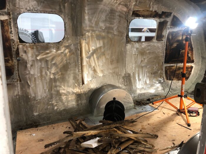 Interiör av en renoverad vagn med exponerad aluminiumvägg och skräp av gammalt virke på golvet, upplyst av en arbetslampa.