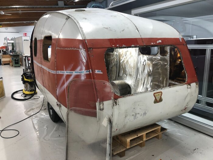 En gammal röd och vit husvagn under renovering i ett verkstadsutrymme, utan fönster och med byggmaterial omkring.