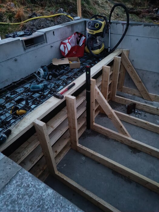 Formar och armering för en betongtrappa i byggprojekt, omgiven av olika verktyg och en dammsugare.