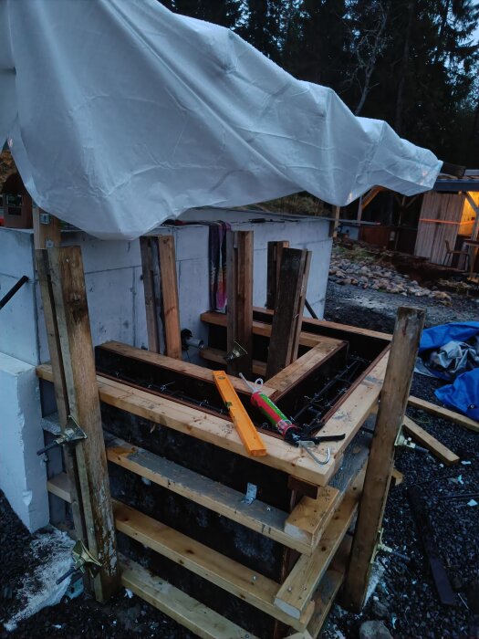 Byggplats med form för betonggjutning, omgiven av trästöd och verktyg. En presenning täcker delvis konstruktionen.