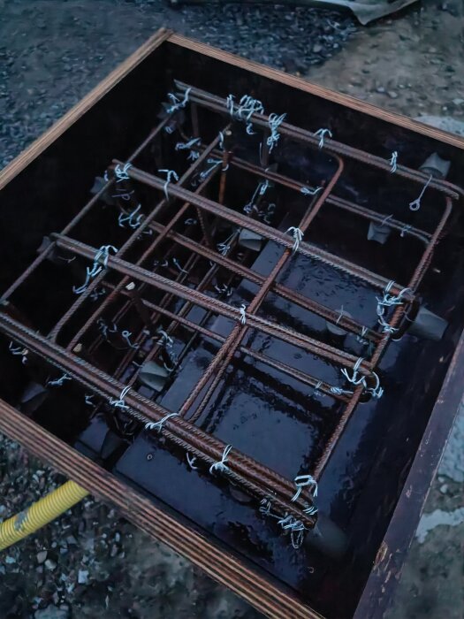 Form med armering av järnstänger och buntband, redo för gjutning, på marken vid ett byggprojekt, omgiven av grus och jord.