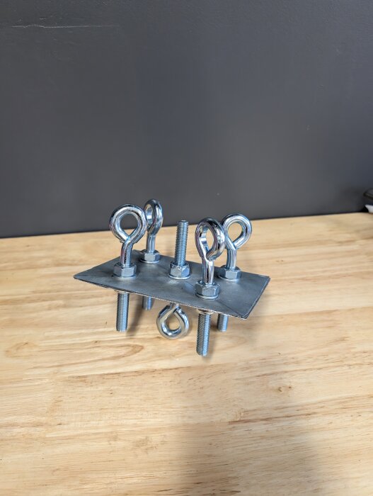 Metallplatta med flera ögleskruvar monterade, del av verktyg för rengöring av rökkanal, på ett träbord.