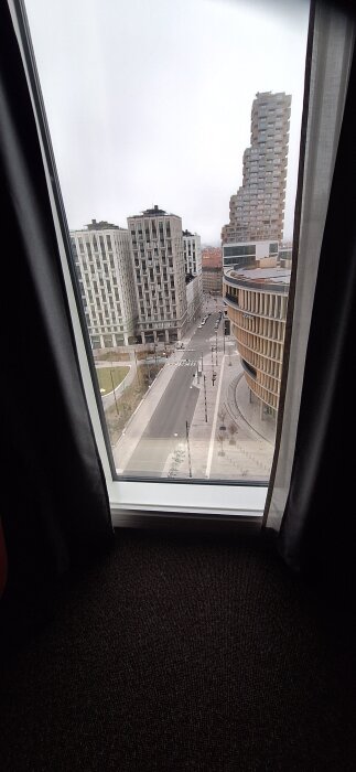 Utsikt från ett fönster över en modern stadsgata med flera höga byggnader och en rundad byggnad till höger.