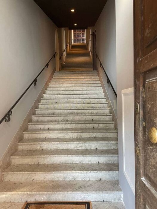 Trappa i ett gammalt renoverat hus från 1920, Rome. Lång vit marmortrappa med svart handledare, leder upp mot ljusöppning.
