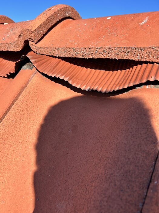 Trasig blykappa på tak med glapp mellan nock och övre pannrad, synlig under klar himmel, potential orsakad av rörelse i takpannorna.