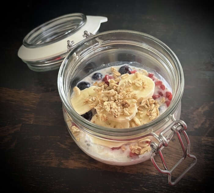 Öppen glasburk med overnight oats toppad med skivad banan, hallon, blåbär och krispiga flingor.
