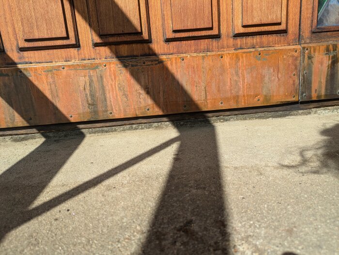 Dörrtröskel i trä med rostiga bleck och skuggor på betonggolv, delvis i dåligt skick där murket trä har avlägsnats, synlig dörr och fönsterkant.