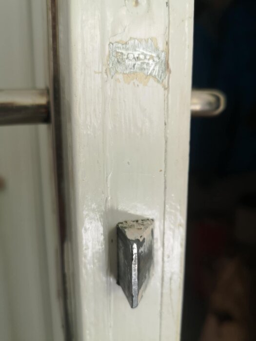 Dörrdetalj med skadad vit färg och metallbeslag, troligen del av lås eller dörrkarm.