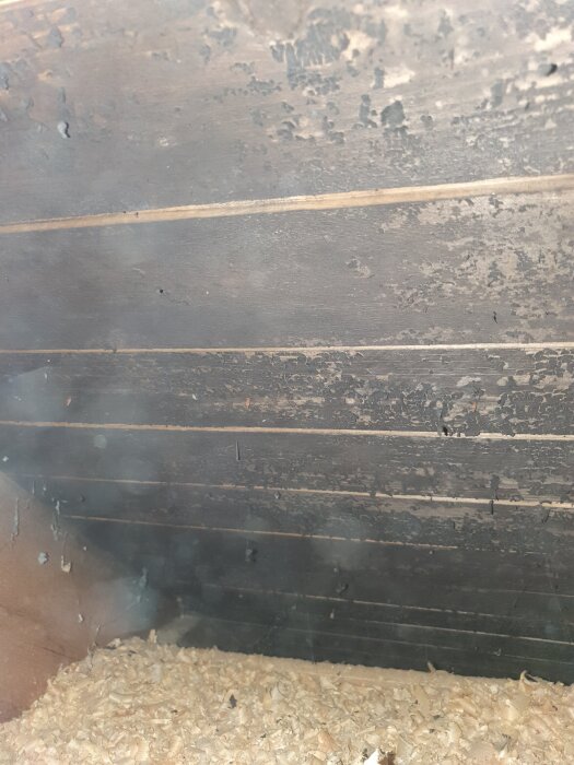 Bilden visar en vind med gamla råspontbrädor som har mörka fläckar, eventuellt mögel, vilka lossnar i fjäll på södersidan.