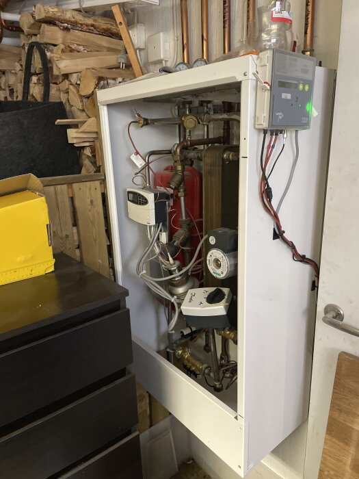 Fjärrvärmecentral med rör och komponenter, inklusive en värmeväxlare och styrenhet, installerad i ett utrymme med trä och verktyg i bakgrunden.