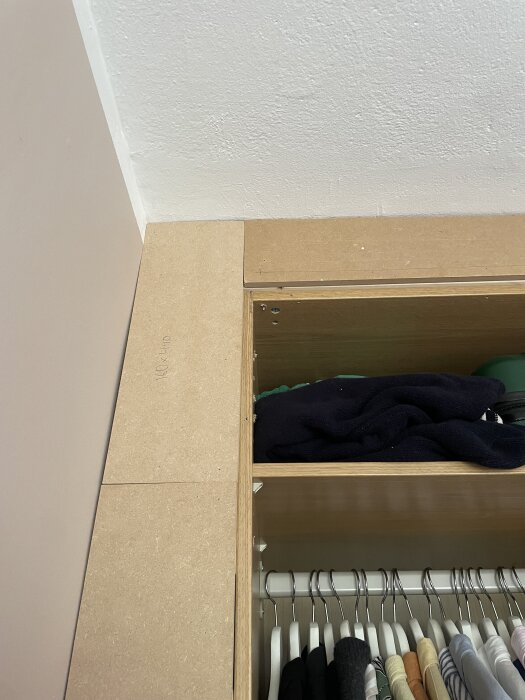Inbyggd garderob med kläder, öppen hylla och klädstång. Ovanpå garderoben en oavslutad träpanel för inbyggnad.