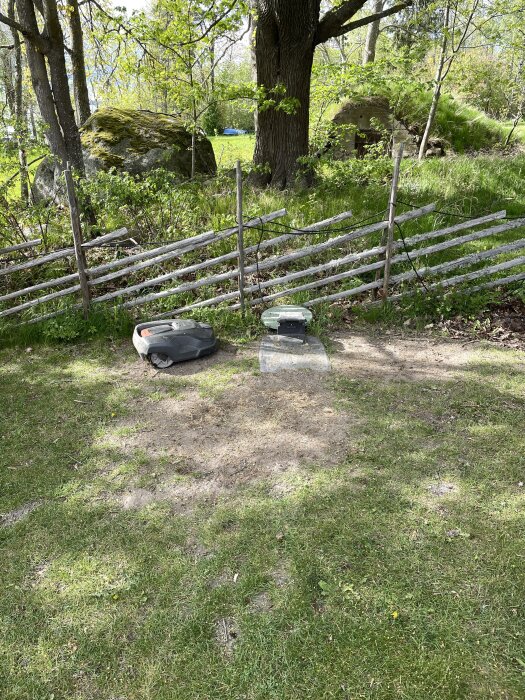 Gräsmatta med en robotgräsklippare nära en laddstation, delvis skymd av ett trästaket. Omkringliggande skogs- och naturmiljö syns i bakgrunden.