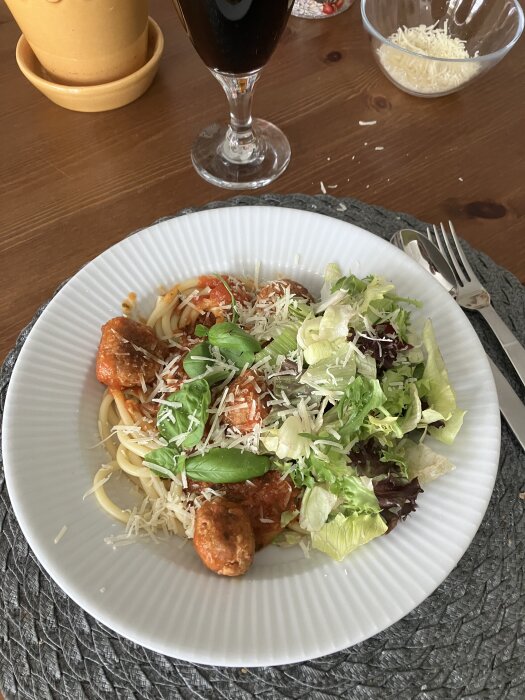 Köttbullar i tomatsås med bucatini och sallad på en tallrik, serverat med ett glas mörk dryck.
