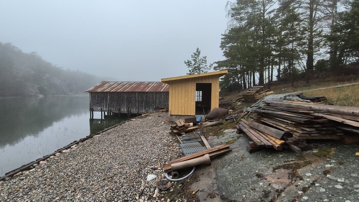 Strandnära bastubyggnation under uppförande med staplade träplankor framför, belägen vid sjö och omgiven av tallar och berg.