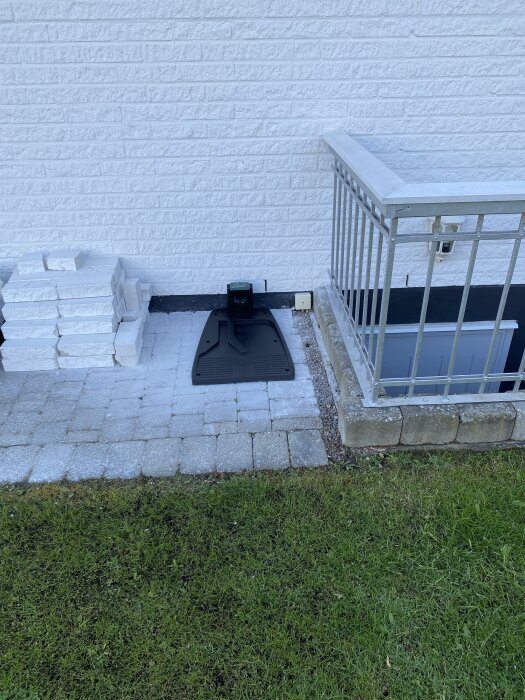 Robotgräsklipparens laddstation på plattbelagd yta intill vit tegelvägg och metallräcke, med staplade plattor i bakgrunden.