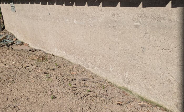 Sliten 80-talssockel med sprickor och saltavlagringar, gränsande till jordig mark och lösa föremål. Ska renoveras med slammat ytskikt.
