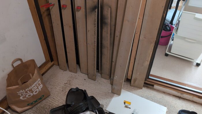 Borttagna plankor står lutade mot väggen, en papperspåse och verktyg på golvet. Del av renoveringsprojekt.