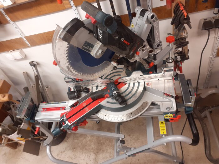 Kap- och gersåg på stativ i verkstad med sågklinga och inställningsrattar, omgiven av olika verktyg och tillbehör.