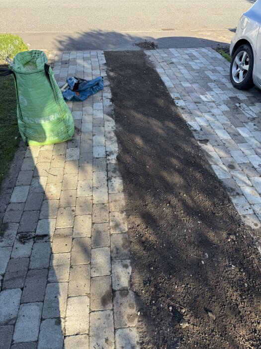 Nyjordad yta på stenlagd uppfart med säck och verktyg i bakgrunden, del av projekt för att odla gräs med Instagramtrick.