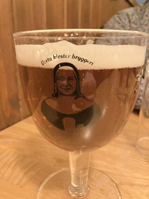 Ölglas med skummig brun dryck, texten "Vreta Kloster bryggeri" och bild av en person på glaset, placerat på träbord mot träpanel.