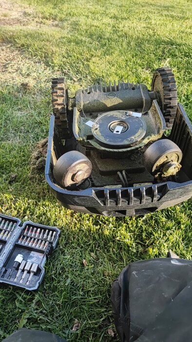 Gräsklipparens undersida med smutsiga knivblad, verktygslåda bredvid för knivbyte på gräsmattan i solljus.