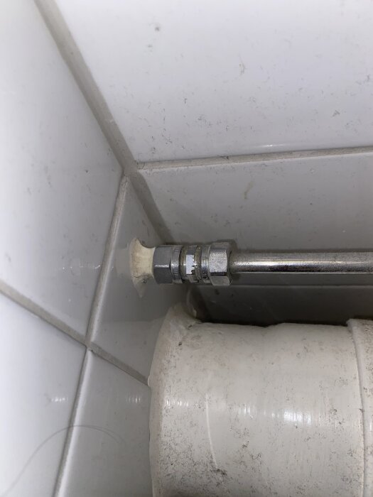Vattenrörsanslutning i badrumshörn, kromrör och vit PVC-rördel, potentiellt för anslutning till WC-stol.