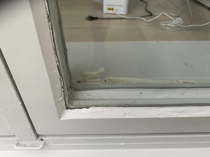 Närbild av ett fönster med synligt sprucket kitt och ojämn målning på karmarna. Inomhusmiljö med sladdar i bakgrunden.