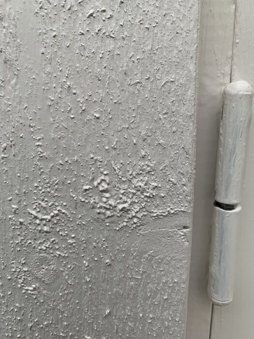 Närbild av en målad träyta på en byggnad, med ojämn och bubblig målning bredvid en gångjärn.