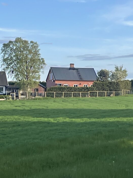 En röd tegelvilla med svart tak och stora fönster, omgiven av gröna fält och ett trästaket i förgrunden.