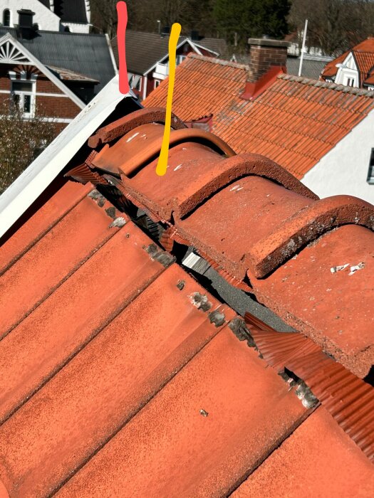 Tegeltak där pannornas toppar inte linjerar med plåtens spets. Rött och orange tegel med synlig åsavslutning och omgivande tak i bakgrunden.