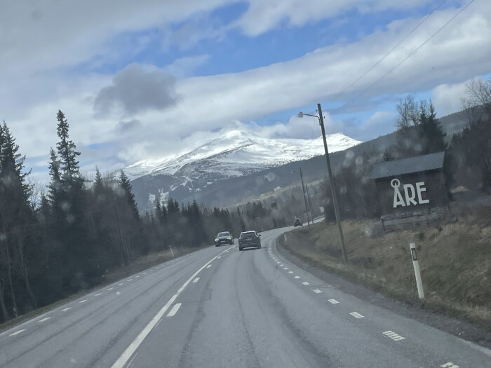 Väg genom skog mot snötäckta berg med skylt som visar "ÅRE" på sidan och bilar som kör mot bergen.