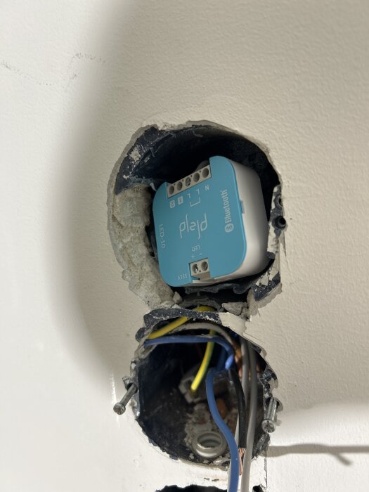 Elektrisk dosa i tegelvägg med synliga elkablar och blå enhet; installation är snett monterad och omgiven av putsad vägg.