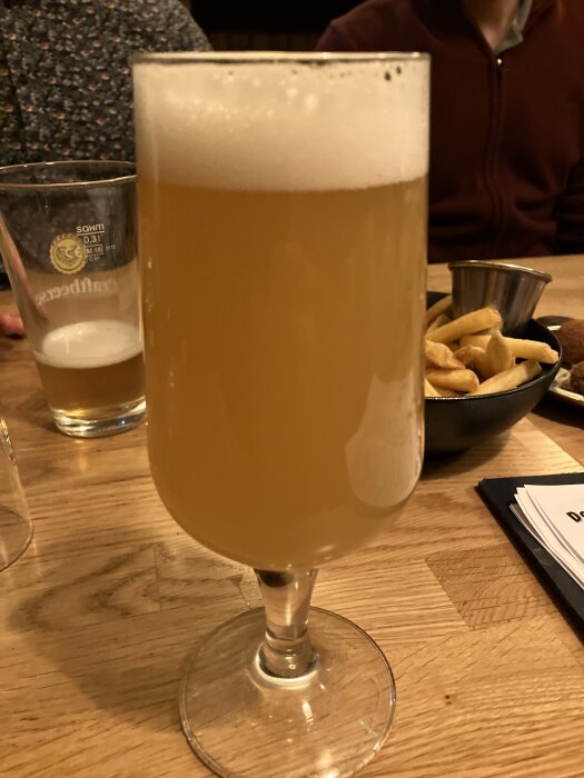 Ett glas med ljus öl med skum på toppen står på ett träbord. I bakgrunden syns en skål med pommes frites och en drinkmeny.