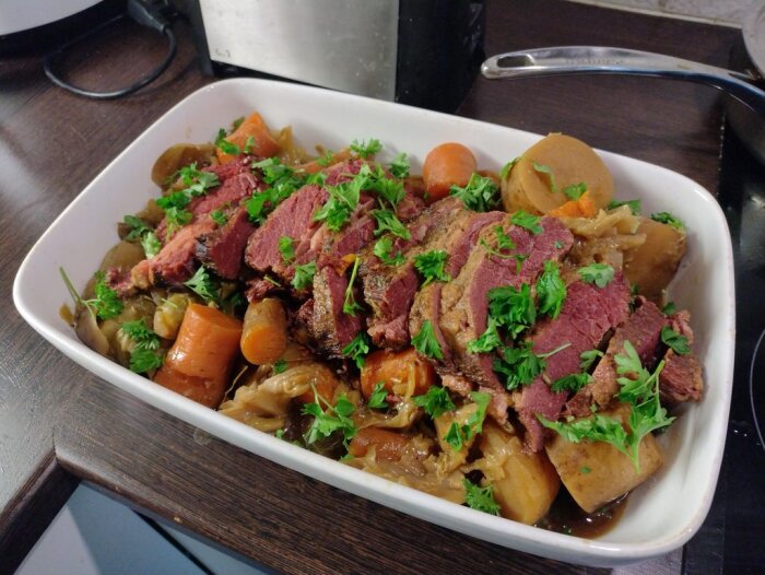 Corned beef med kål och korv i en vit ugnsform, dekorerad med persilja, förberedd i slow cooker.