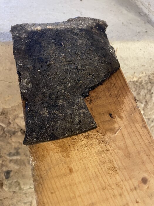 Svart mattliknande material på träplanka, misstänkt vara asfalt eller bitumen, med uppvikta kanter.