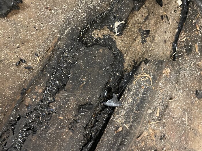 Svart mattliknande material under ett borttaget trägolv, misstänkt asfalt eller bitumen, med omgivande smuts och träflisor i en källare från 30-talet.