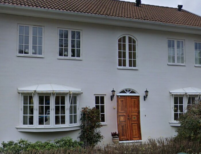 Vitputsat hus med bruna trädetaljer, tak av tegel, flera fönster och en stor träport. Förgrund med grönska och buskage.