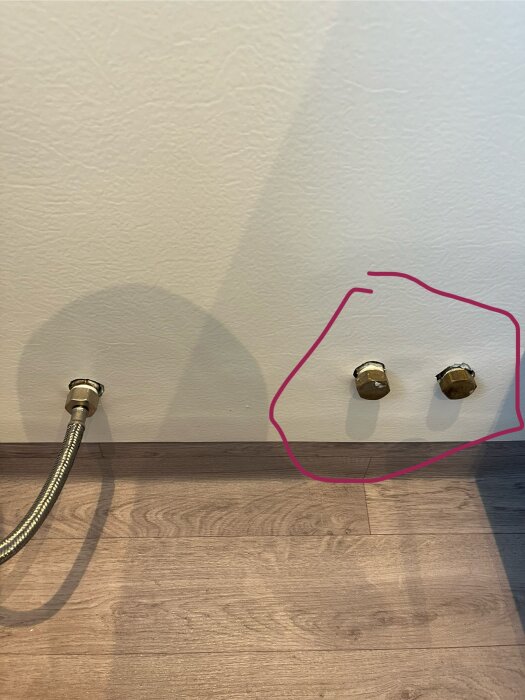 Vägg med två oanvända vattenrör täckta med metallpluggar; ena röret är markerat med en rosa cirkel.