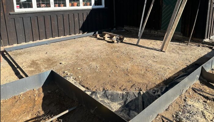 Uteplats under konstruktion med naturgrus som bärlager, omgärdat av kanter i cortenstål vid husfasaden. Mark är ännu ej vibrerad.