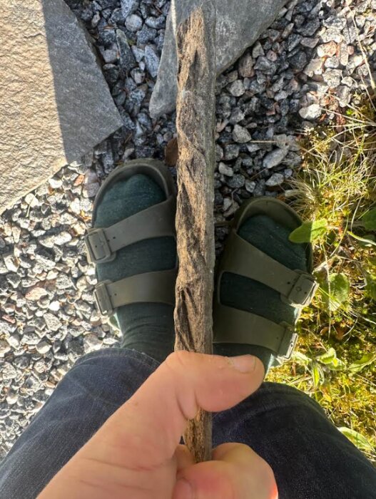 En hand jämför tjockleken på en skifferhäll mot en fot i sandal, omgiven av grus och gräs.
