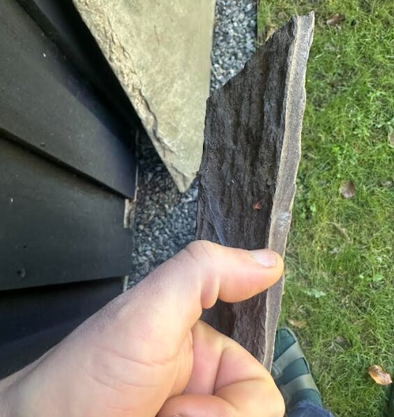 Hand som håller en tunn skifferhäll mot en bakgrund av gräs och grusbeläggning.