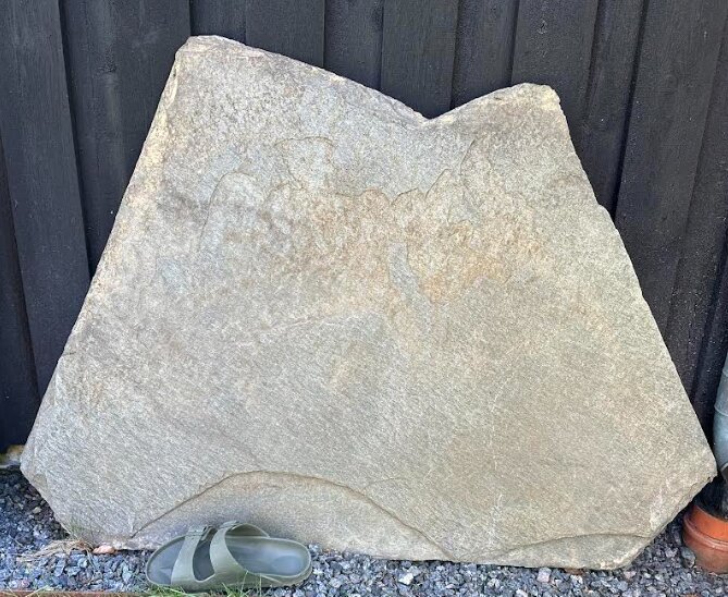 En stor, oregelbunden skifferhäll lutad mot en mörk vägg, med en toffel placerad framför för att visa storleken.