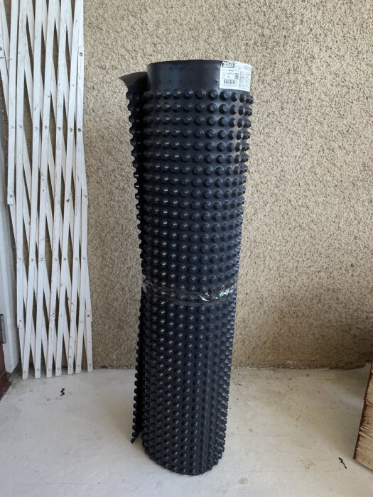 Rulle av svart plast med knottrig yta, används som fuktskydd för väggar och golv, stående mot en vägg i ett rum.