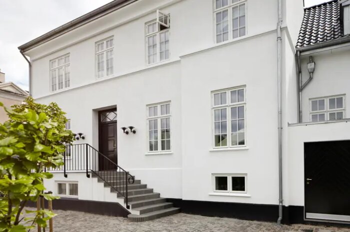 Vit putsad fasad på tvåvåningshus med trappa och svart dörr, omgiven av gröna växter och kullersten.