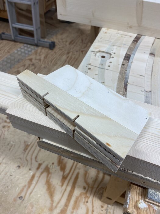 Träbitar placerade i vinkel för att fräsa ur hål med hjälp av en mall, visande en arbetsyta av trä och golv av plywood i en verkstad.