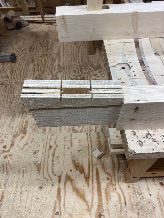Träbitar med frästa hål för kilar, ligger på arbetsbänk med golv av plywood i bakgrunden.