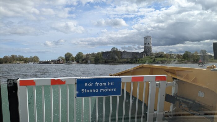 Färja med skylt "Kör fram hit, Stanna motorn" på vattnet med medeltida fästning och torn i bakgrunden under molnig himmel.