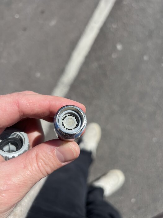 Hand håller en Gardena-koppling med en vit plastplugg inuti, som troligen begränsar vattenflödet. Bakgrund av asfalt.