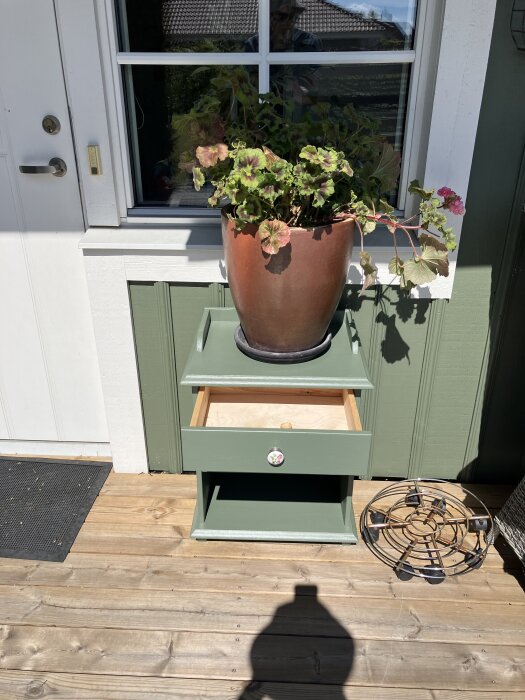 Nymålad grön hurts med en öppen låda, en blomkruka på toppen, placerad utomhus nära en vit dörr och ett fönster.